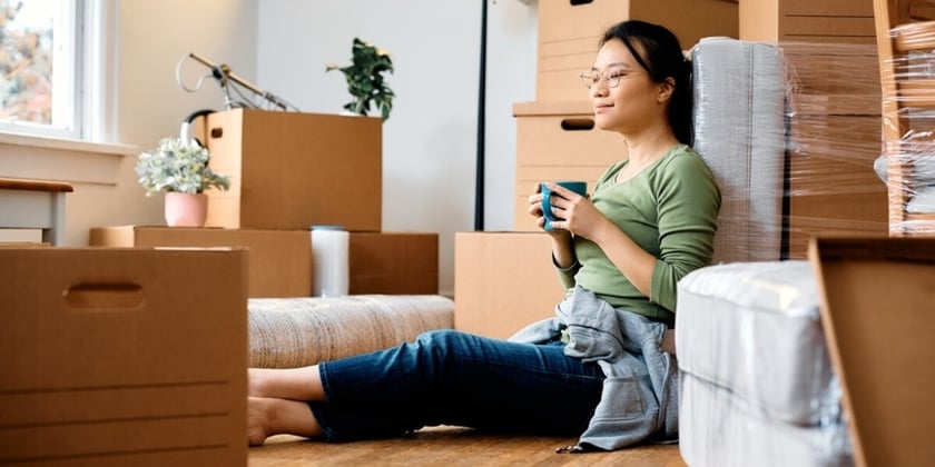 Young Asian woman sitting on the floor among cardboard boxes while moving into new apartment