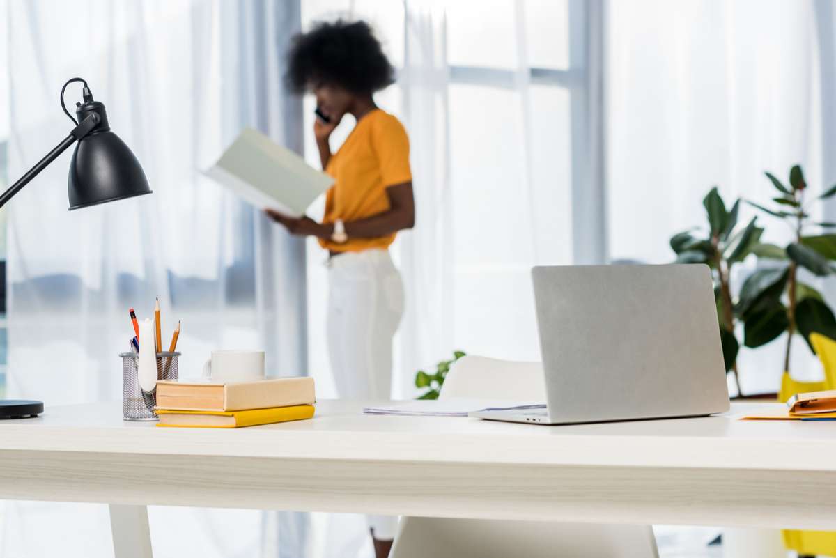 Selective focus of laptop at workplace and african american freelancer talking on smartphone on background at home (R) (S)