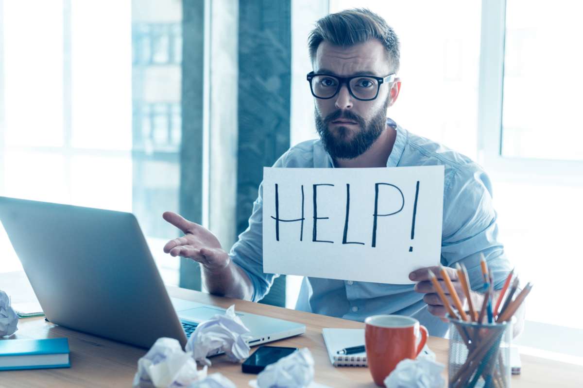 Frustrated young beard man holding piece of paper and asking for help while sitting at his working place in office